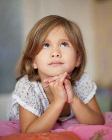 female child in nightgown kneeling at her bed with clasped hands under chin while looking up