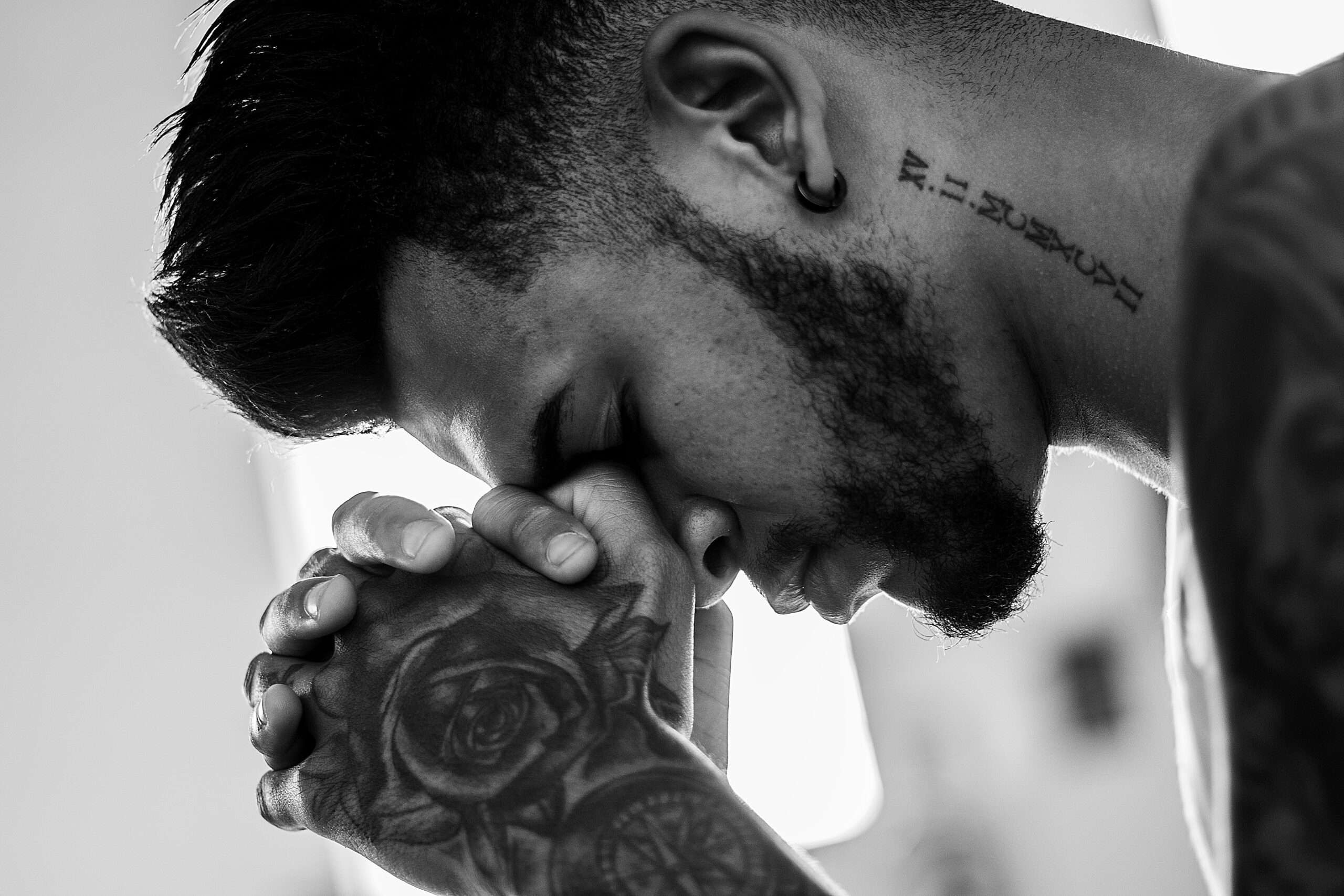 man with tattoos and black hair and beard praying with head resting on hands in a fist